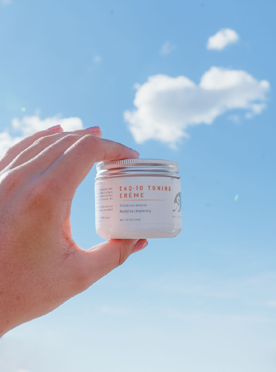 Woman's hand holding a jar of White Egret's CoQ-10 Toning Creme up to a blue sky with white clouds
