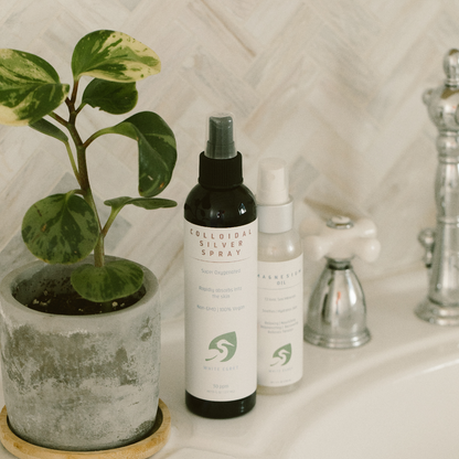 White Egret's Colloidal Silver Spray on a bathroom counter next to White Egret's Magnesium Oil with a potted plant beside it