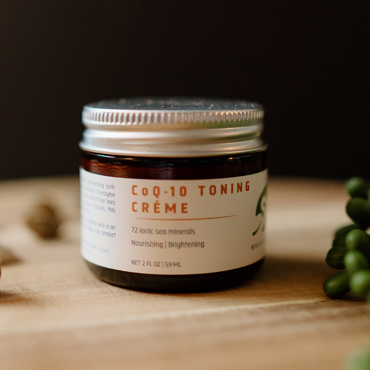 White Egret's CoQ-10 Toning Creme in a glass jar on a cutting board with pebbles and fake plants placed around the jar